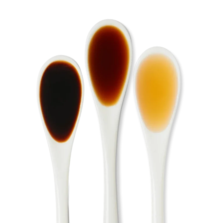 Three white spoons hold different liquids: dark brown, medium brown, and light amber, displayed side by side on a white background.
