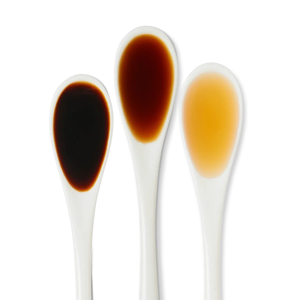 Three white spoons hold different liquids: dark brown, medium brown, and light amber, displayed side by side on a white background.