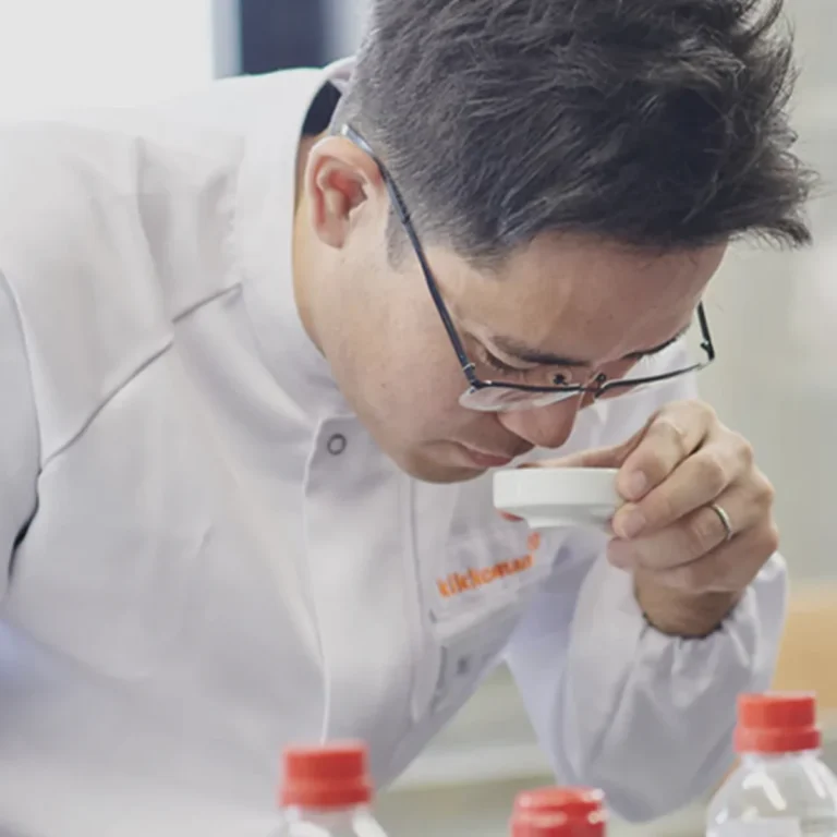 A person in a lab coat and glasses closely smelling the contents of a small white dish, with bottles visible on the table in front of them.