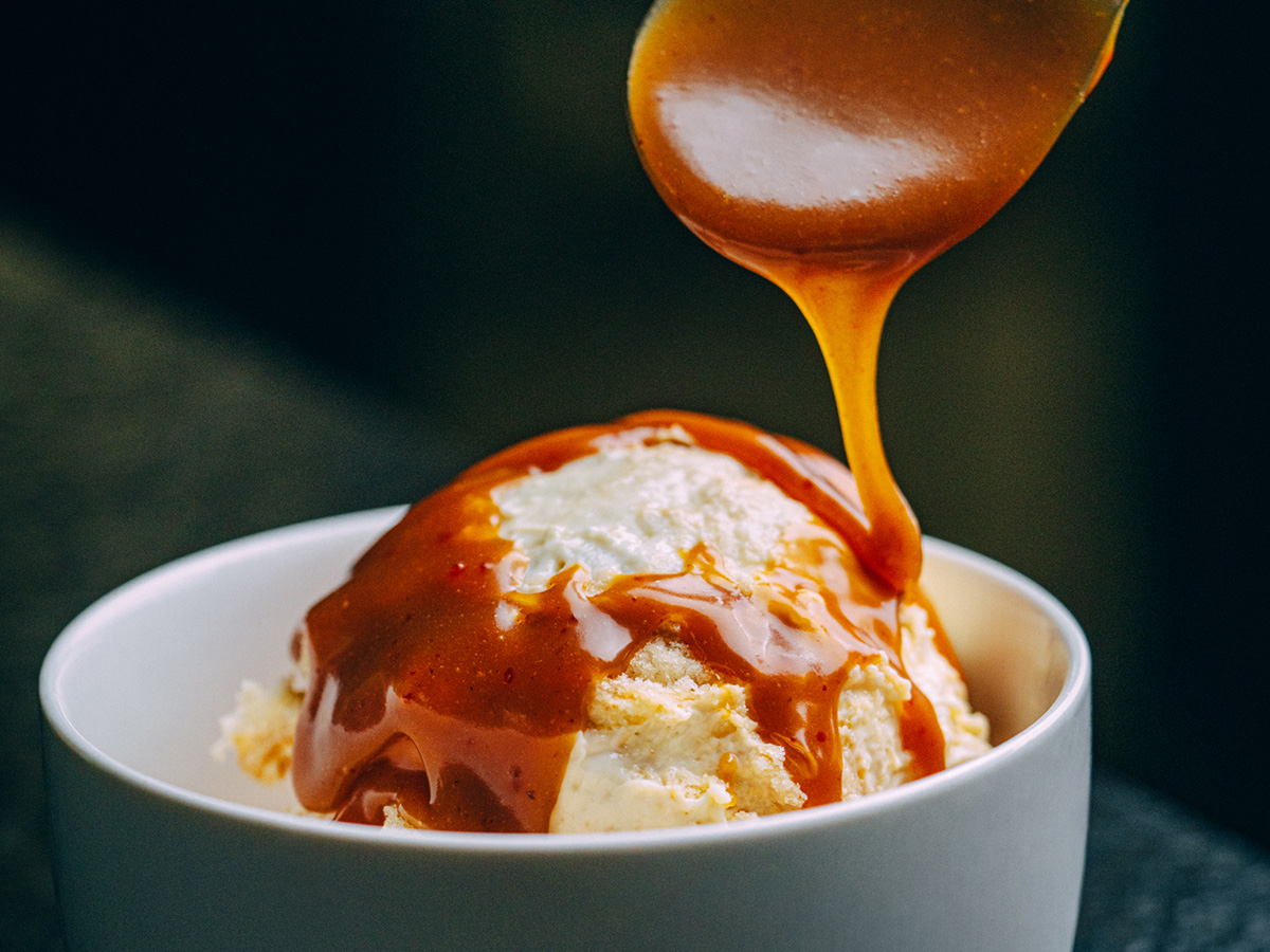 A spoon pours caramel sauce over a scoop of vanilla ice cream in a white bowl.