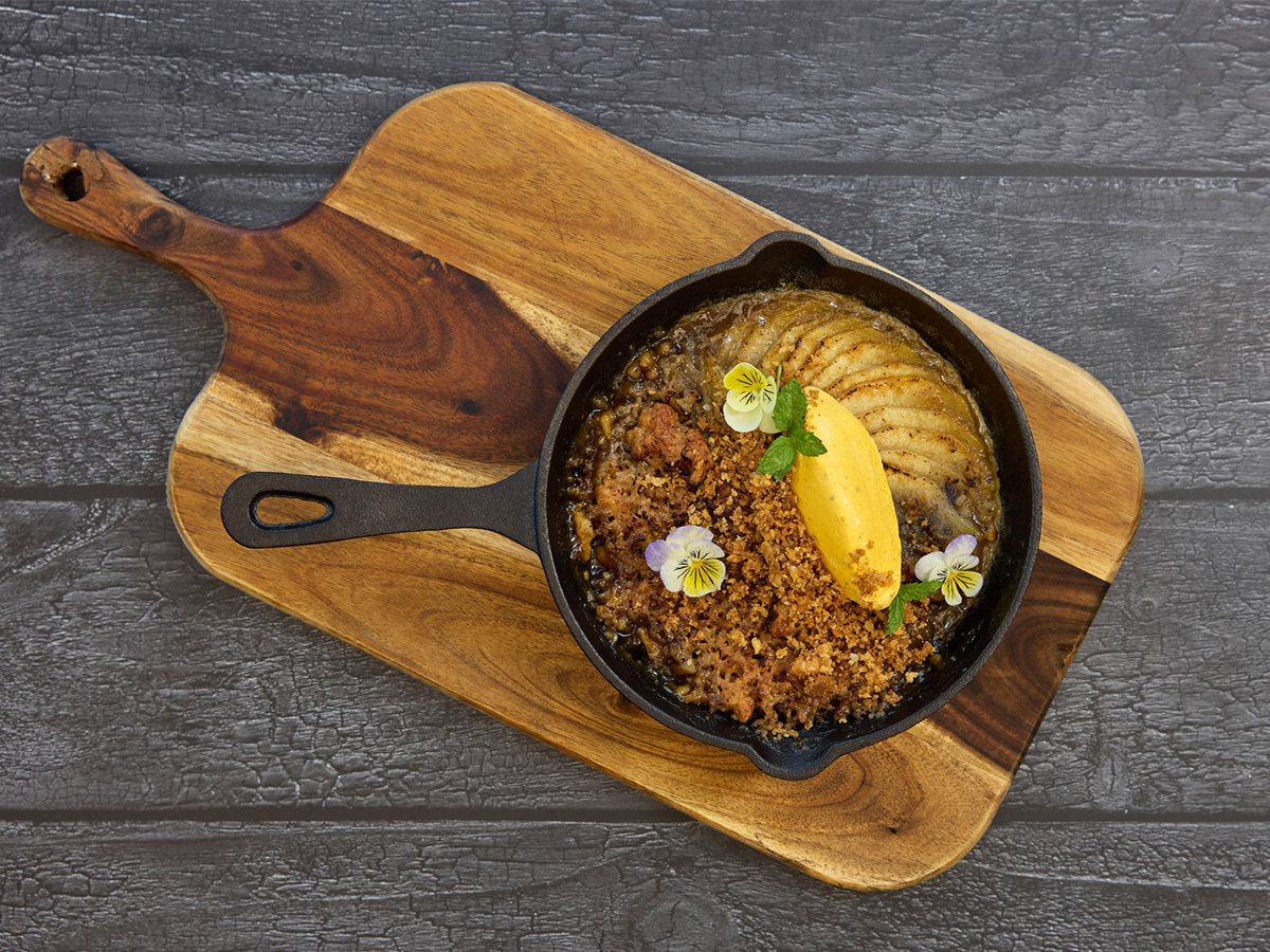 A small cast iron skillet with a baked dessert topped with a scoop of yellow ice cream, edible flowers, and mint, placed on a wooden serving board.
