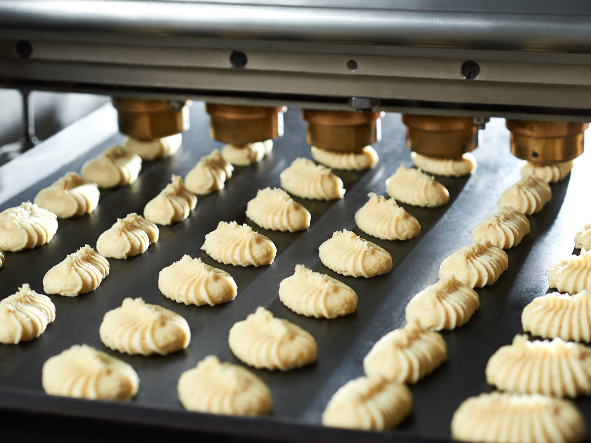 Rows of unbaked cookies are being shaped by a machine onto a baking tray in a commercial bakery setting.