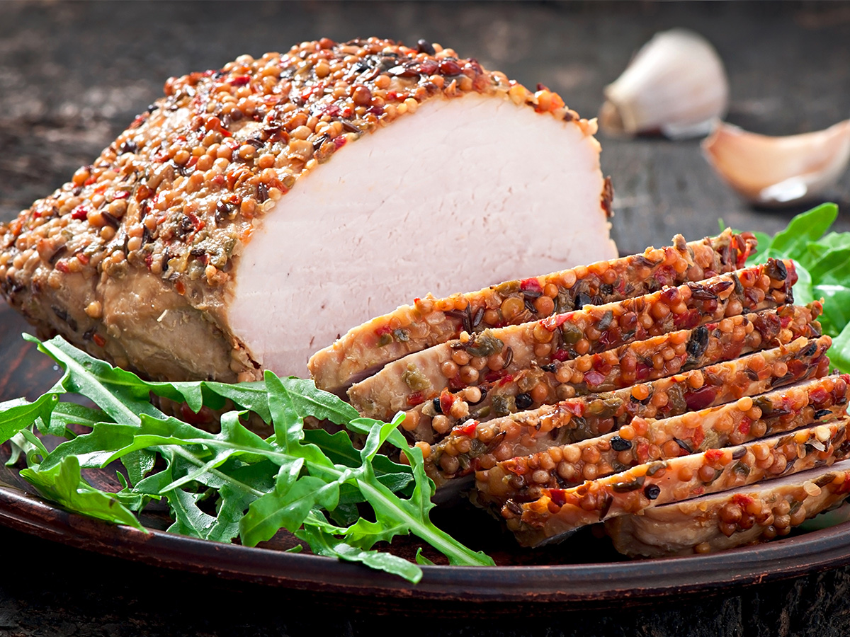 A sliced roast pork loin coated with whole grain mustard and pepper, served on a plate with fresh arugula leaves.