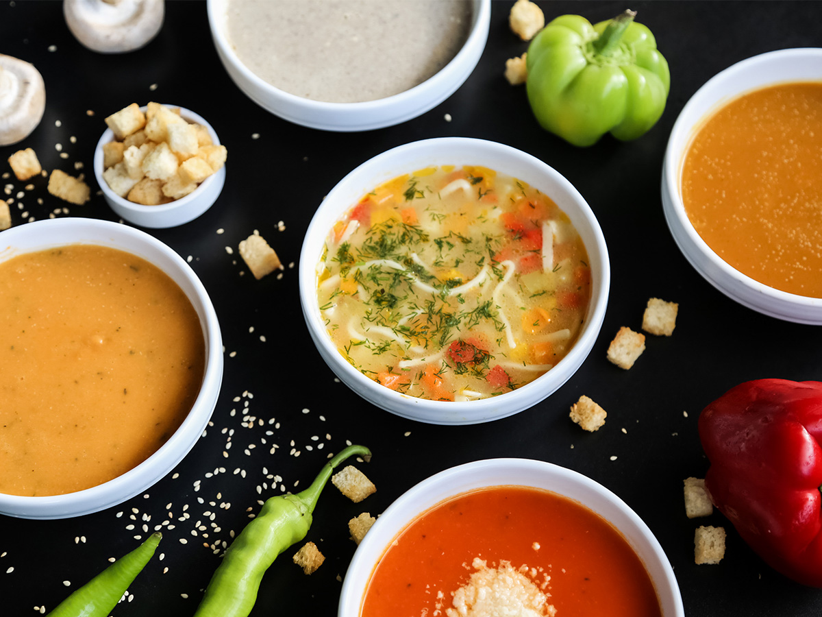 Assorted bowls of soup with various vegetables, croutons, and seasonings arranged on a black surface.