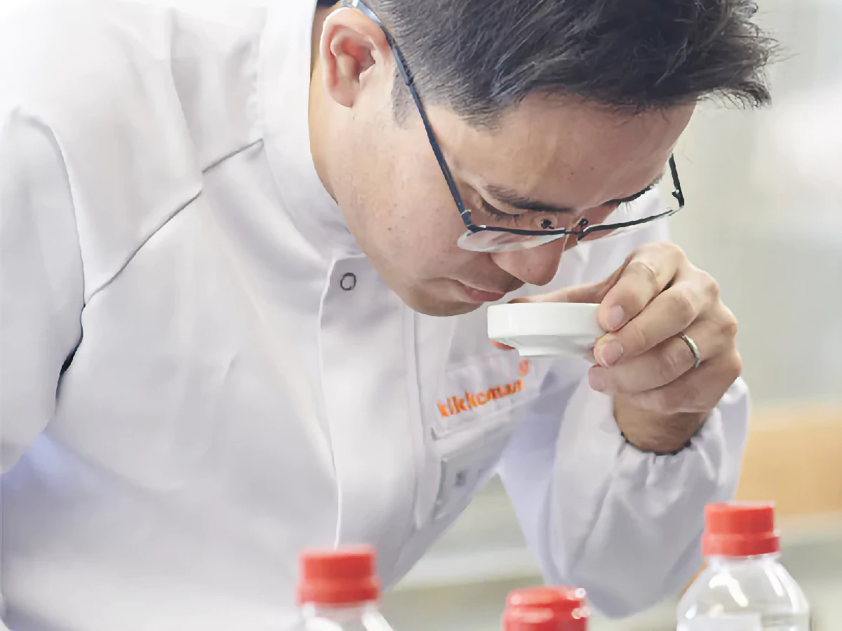 A person in a white lab coat and glasses is smelling the contents of a small white cup, with several bottles with red caps visible in the foreground.