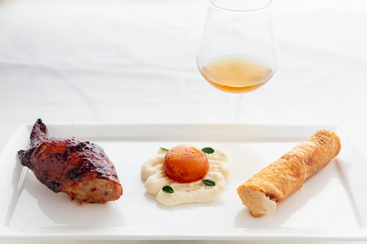 A plated dish with a piece of roasted meat, a round orange garnish on a white puree with herbs, a rolled pastry, and a glass of amber liquid.