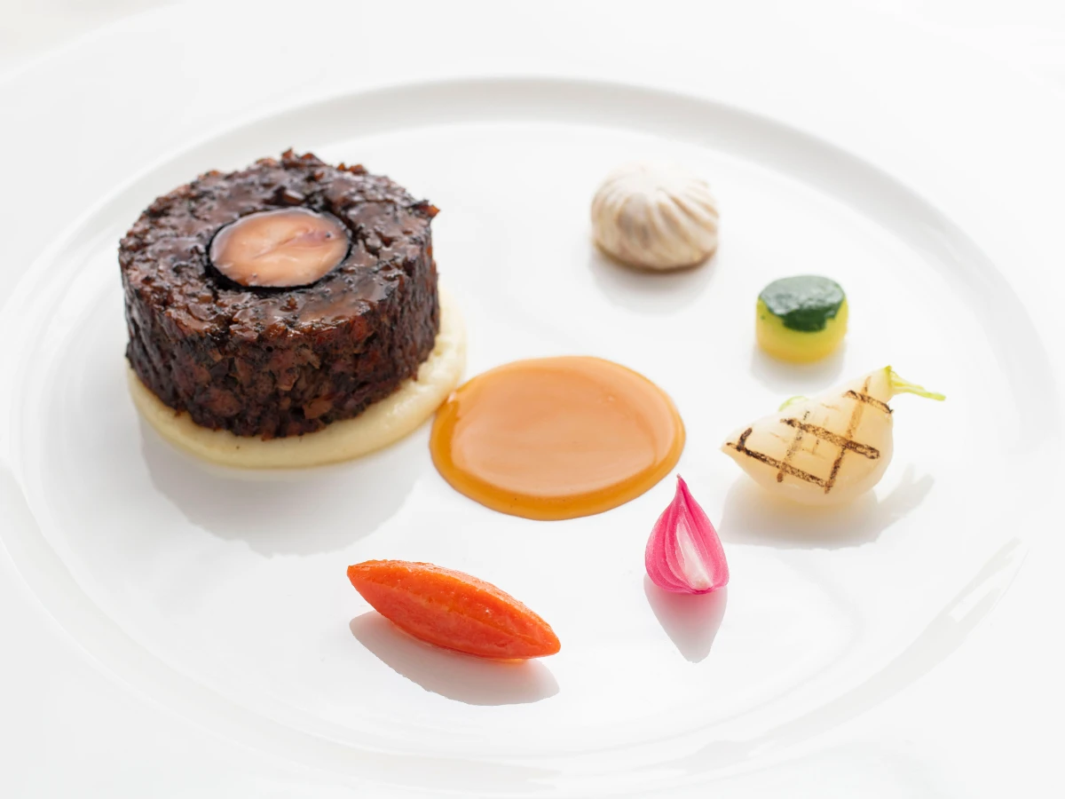 A gourmet dish with a circular meat medallion on puree, surrounded by colorful vegetable garnishes, sauce, and a small piped cream portion, artfully arranged on a white plate.