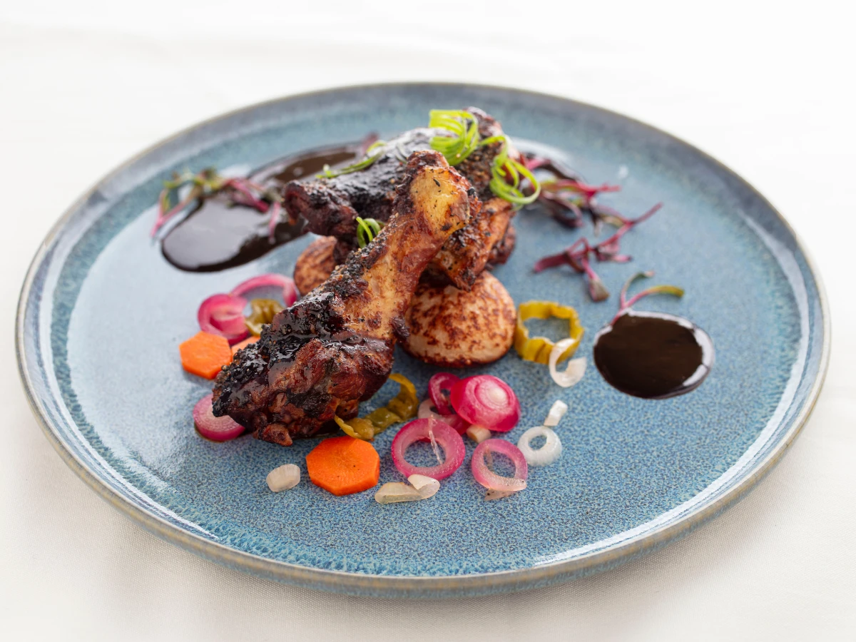 A blue plate with grilled chicken drumsticks on roasted potatoes, garnished with sliced onions, carrots, and green herbs, and drizzled with dark sauce.