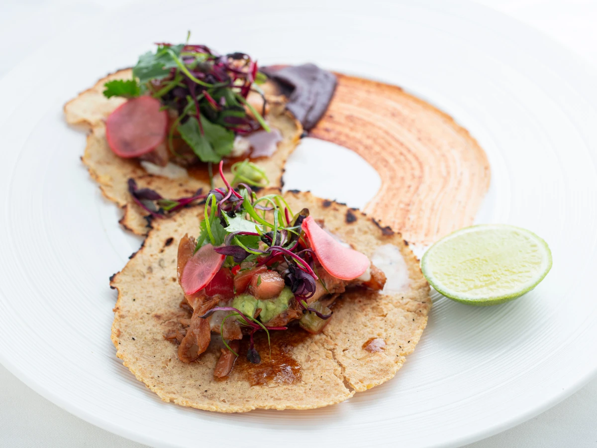 Two gourmet tacos topped with greens, sliced radish, and vegetables on a white plate, with a smear of sauce and a lime wedge on the side.