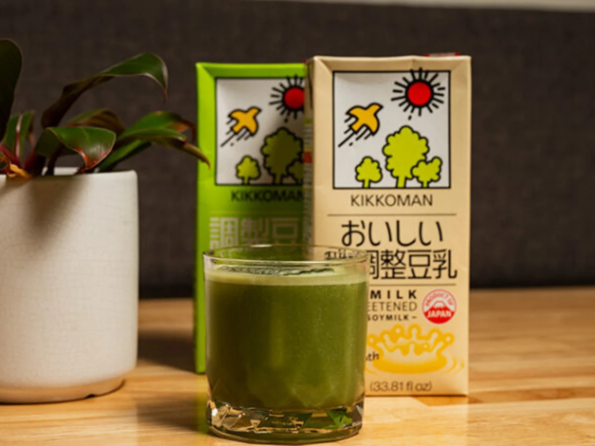 A glass of green soy milk sits on a wooden table in front of two Kikkoman soy milk cartons, with a potted plant on the left.