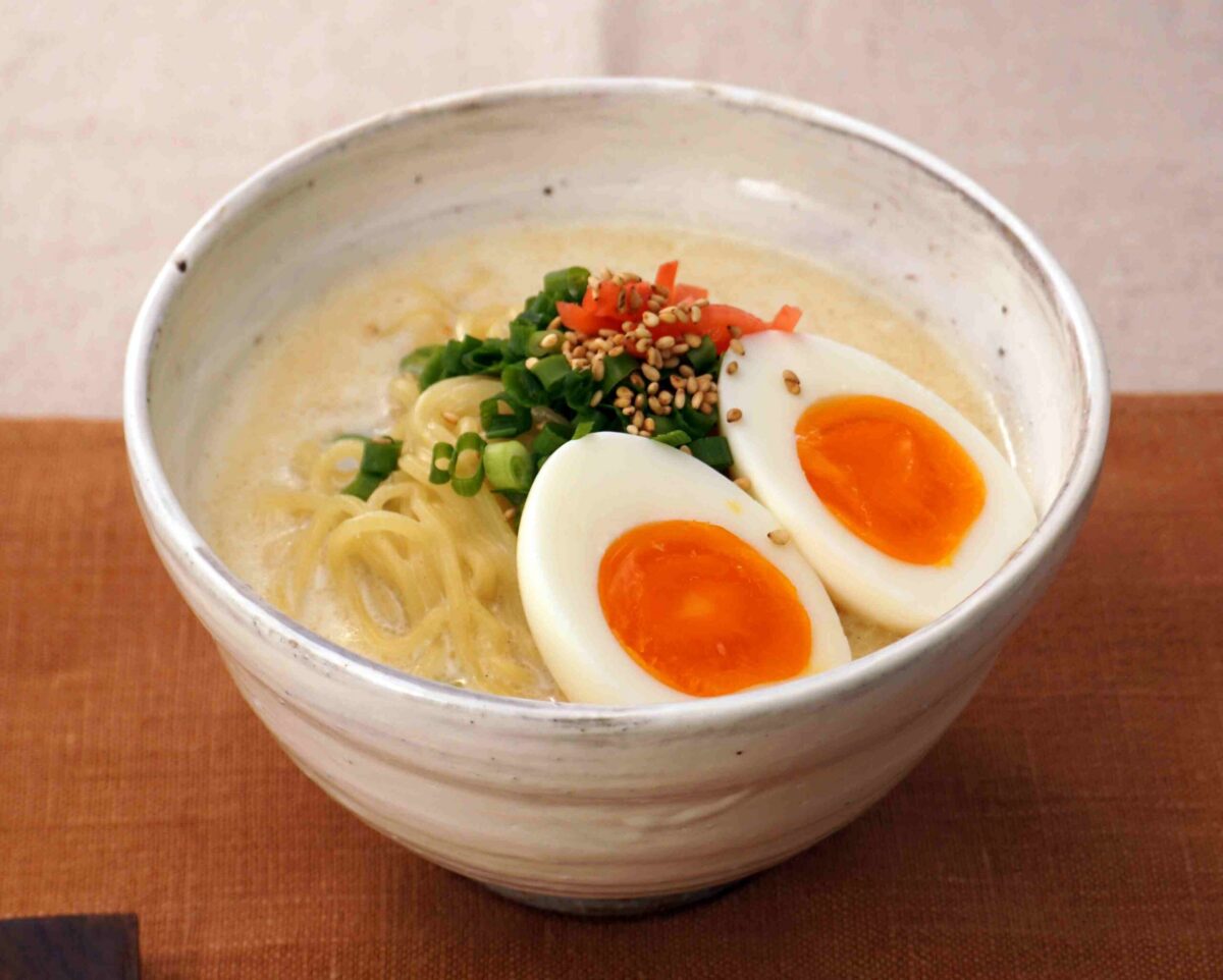 A bowl of ramen with sliced soft-boiled eggs, chopped green onions, sesame seeds, and diced red garnish on top.