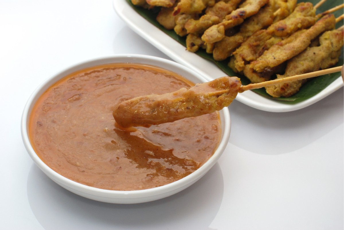 A skewer of grilled meat is dipped into a bowl of peanut sauce, with more skewers on a plate in the background.
