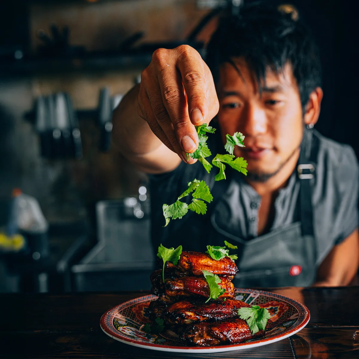 A person garnishes a plate of cooked chicken wings with fresh cilantro in a kitchen setting.