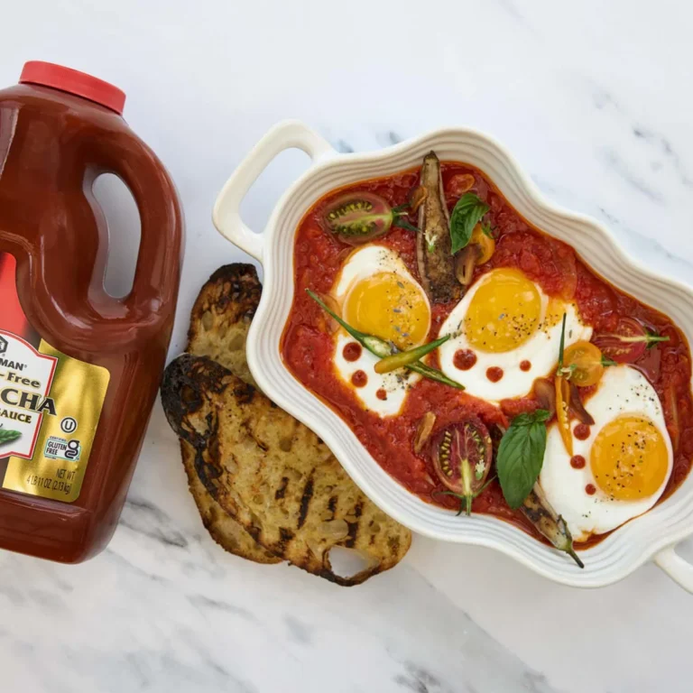 A baked egg dish with tomato sauce and vegetables in a white dish, served with grilled bread and a bottle of sriracha sauce on a white surface.