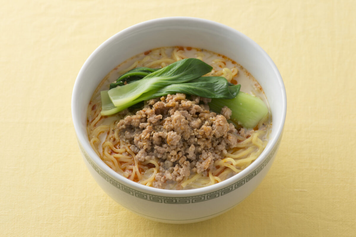 A bowl of noodle soup topped with ground meat and bok choy, served in a white bowl on a yellow surface.