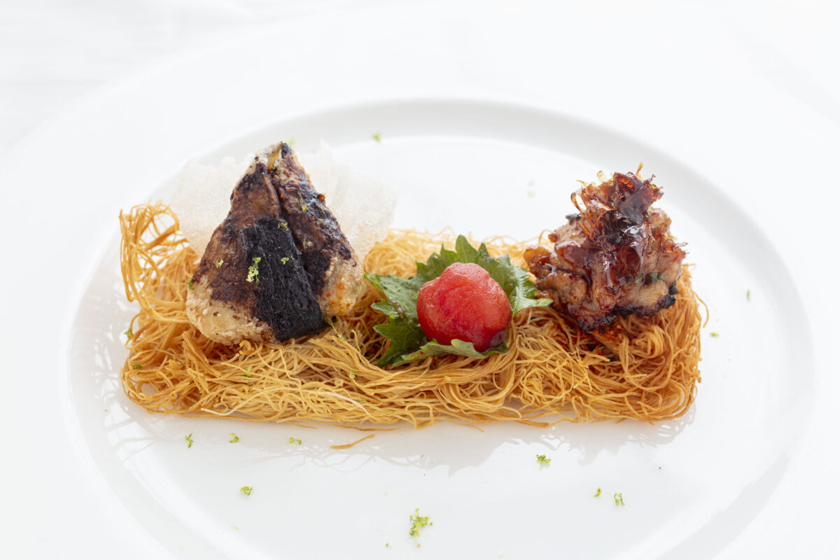 A plated dish features a grilled onigiri, a nest of crispy noodles, a small mound of shredded meat, a green shiso leaf, and a small red garnish, all arranged on a white plate.