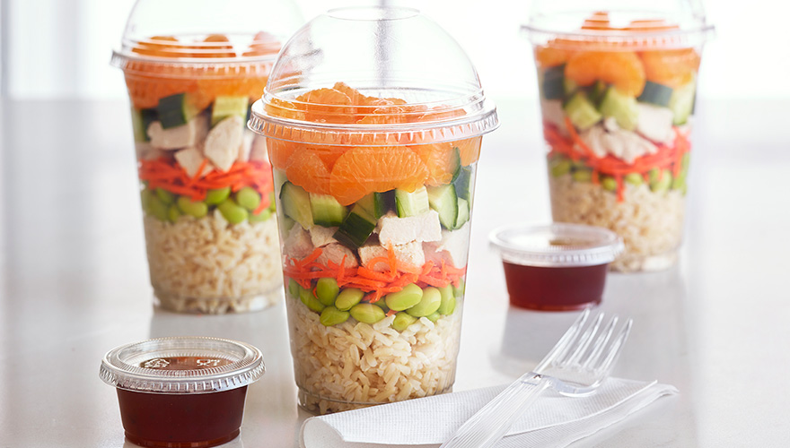 Clear plastic cups filled with layers of brown rice, edamame, carrots, cucumbers, chicken, and mandarin orange slices, with small containers of dressing and plastic cutlery on the side.