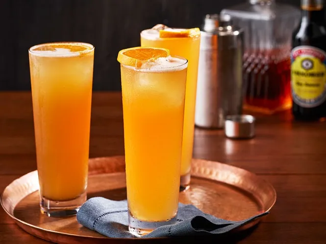 Three tall glasses of an orange-yellow cocktail with orange slices on a copper tray, with a cocktail shaker, bitters, and a decanter in the background.