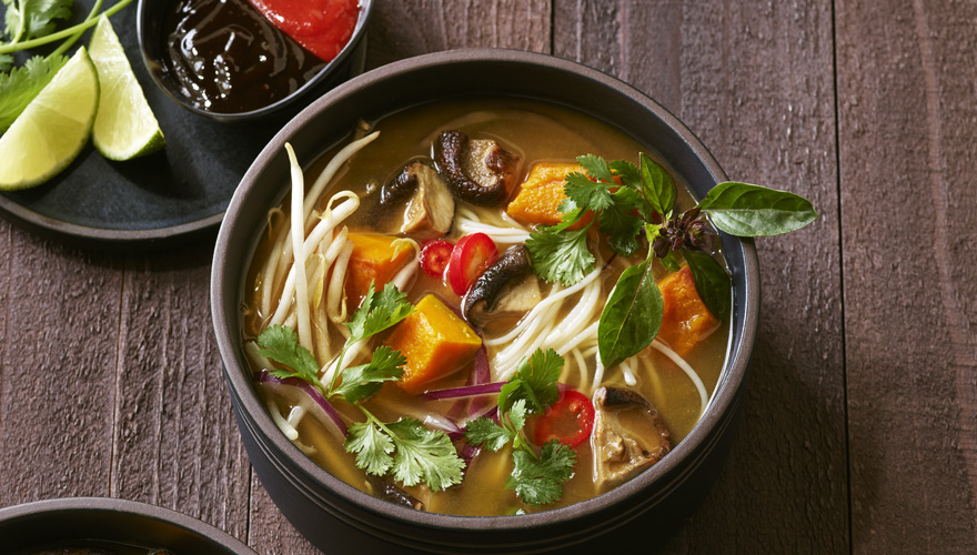 A bowl of soup with noodles, mushrooms, pumpkin, red chili, cilantro, and basil, served on a wooden surface with lime wedges and sauces on the side.