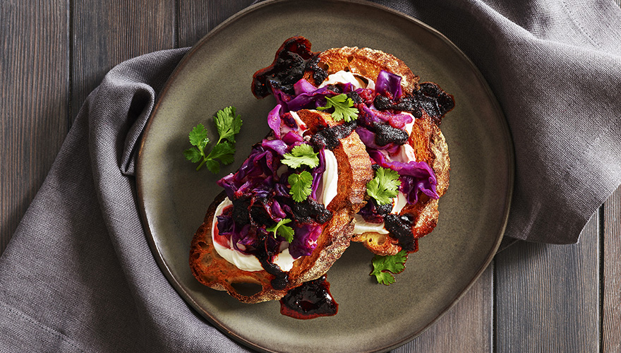 Two slices of toasted bread topped with creamy spread, red cabbage, dark sauce, and fresh cilantro, served on a gray plate with a napkin on a wooden surface.