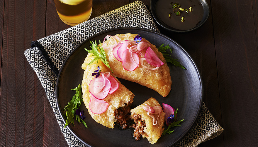 Two golden-brown empanadas filled with ground meat, topped with pickled radish and onions, served on a black plate with greens and edible flowers, next to a dipping sauce.