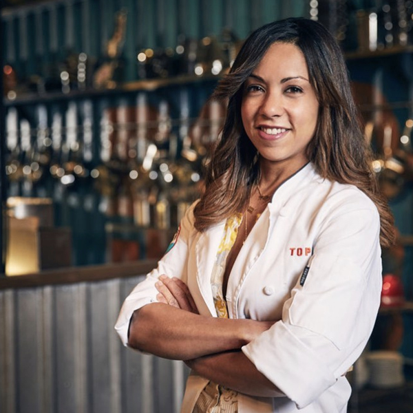 A woman wearing a white chef's coat stands with arms crossed, smiling, in a kitchen with shelves of pots and pans in the background.