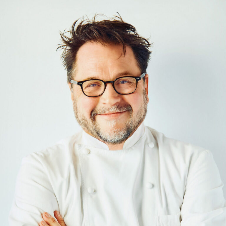 A middle-aged man with glasses and a beard smiles at the camera while wearing a white chef's coat against a plain light background.