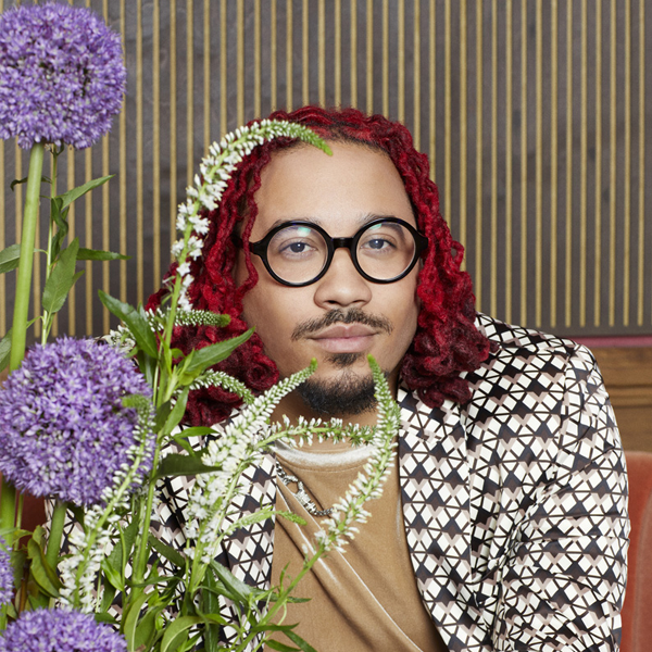A person with red curly hair and round glasses sits behind purple allium flowers, wearing a geometric-patterned jacket and a tan shirt, with a striped background behind them.