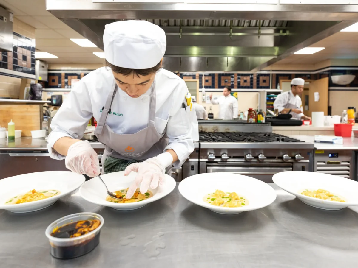 A chef in a white uniform and hat carefully plates pasta dishes in a commercial kitchen, with three other chefs working in the background.