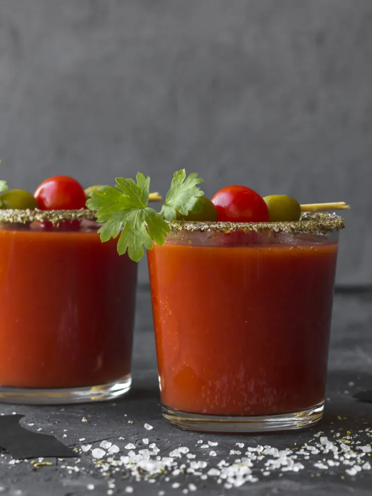 Two glasses of Bloody Mary cocktails, garnished with cherry tomatoes, green olives, and parsley, sit on a dark surface with salt sprinkled nearby.