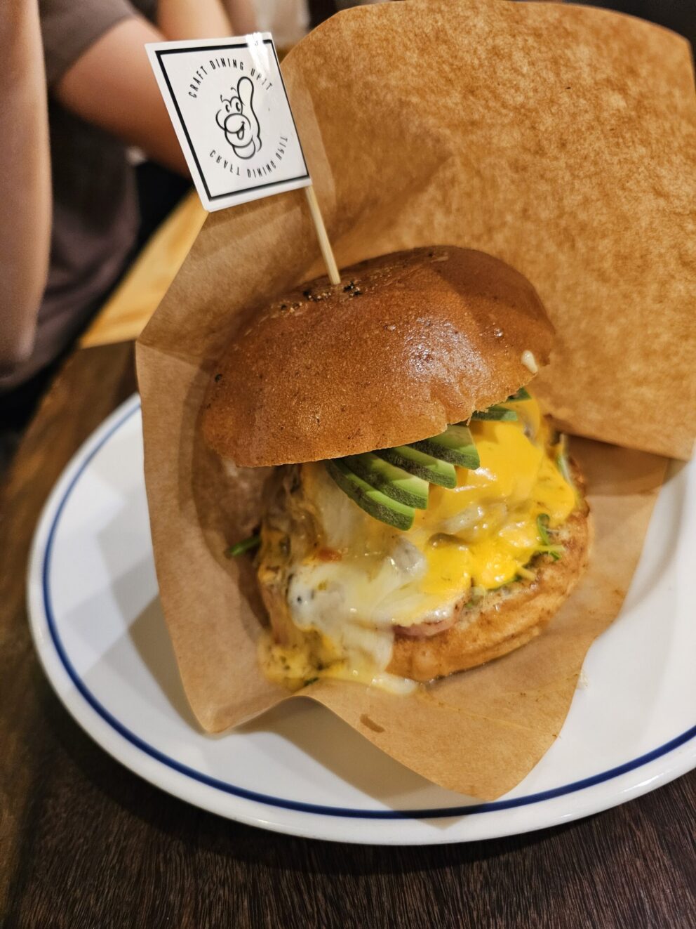 A cheeseburger with melted cheese, sliced avocado, lettuce, and a brown bun, served on a white plate with a small flag on top.