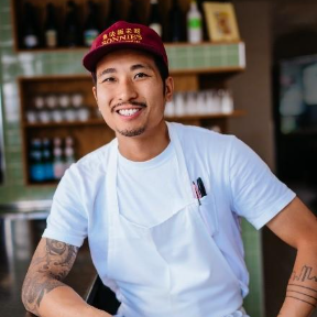 A smiling person wearing a red cap, white t-shirt, and apron stands behind a counter in a restaurant or cafe setting.