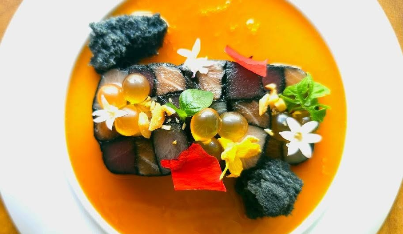 A plated dish with a checkered fish terrine, orange sauce, black crumbs, edible flowers, green herbs, and golden spheres, arranged decoratively on a white plate.