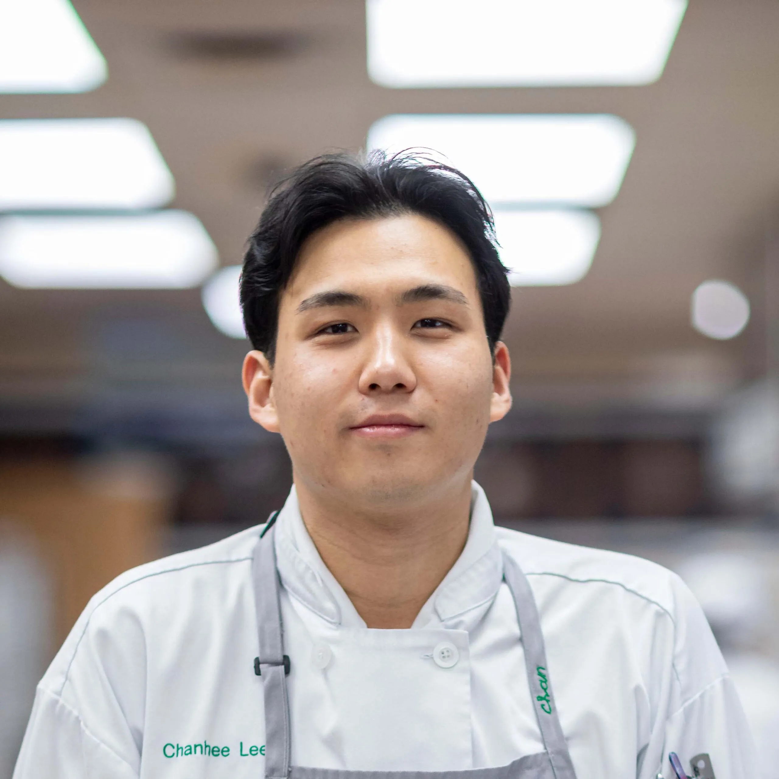 A chef in a white uniform and apron stands indoors, looking at the camera with a neutral expression.