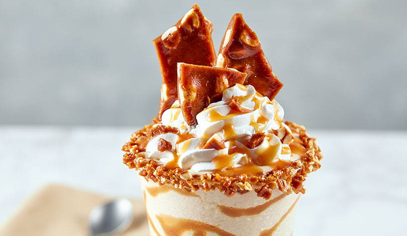A milkshake topped with whipped cream, caramel sauce, and pieces of peanut brittle, with crushed nuts around the rim.