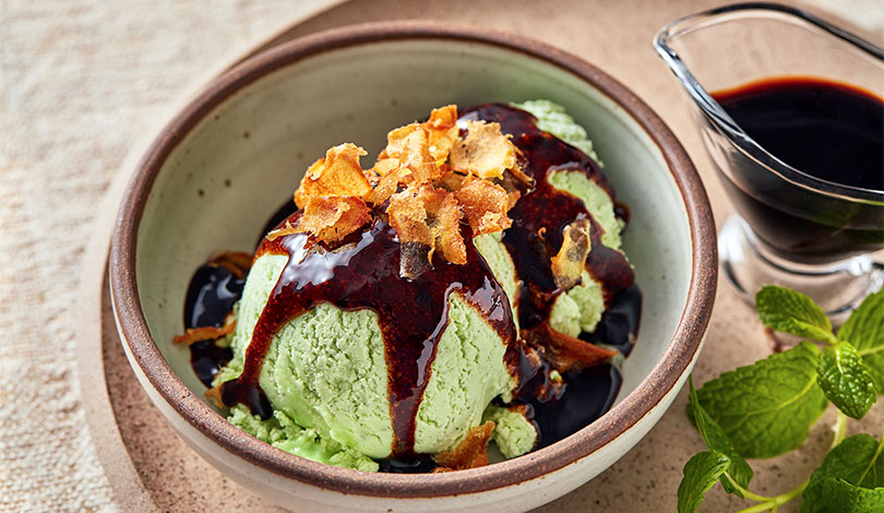 Two scoops of green ice cream topped with dark syrup and crispy flakes in a bowl, with a glass container of syrup and mint leaves beside it.