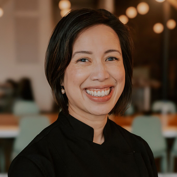 A woman with short dark hair and a black top smiles at the camera in an indoor setting with blurred lights and chairs in the background.
