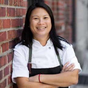 A person wearing a white chef's coat and black apron stands with arms crossed, smiling, in front of a brick wall.