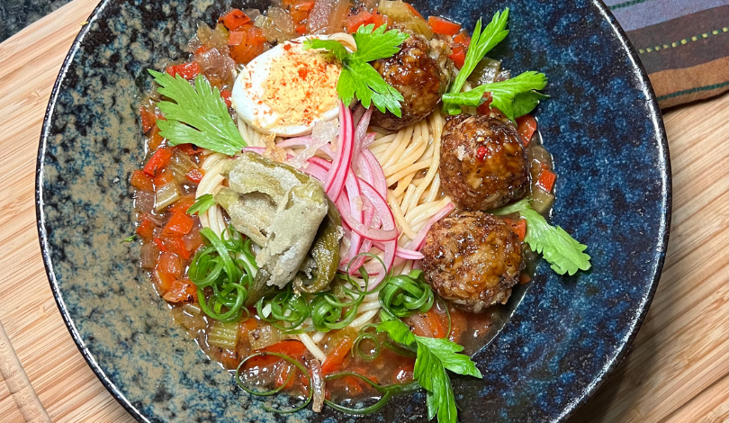 A dark ceramic plate with spaghetti, three meatballs, a soft-boiled egg, pickled onions, sliced peppers, celery leaves, and a chunky vegetable sauce.