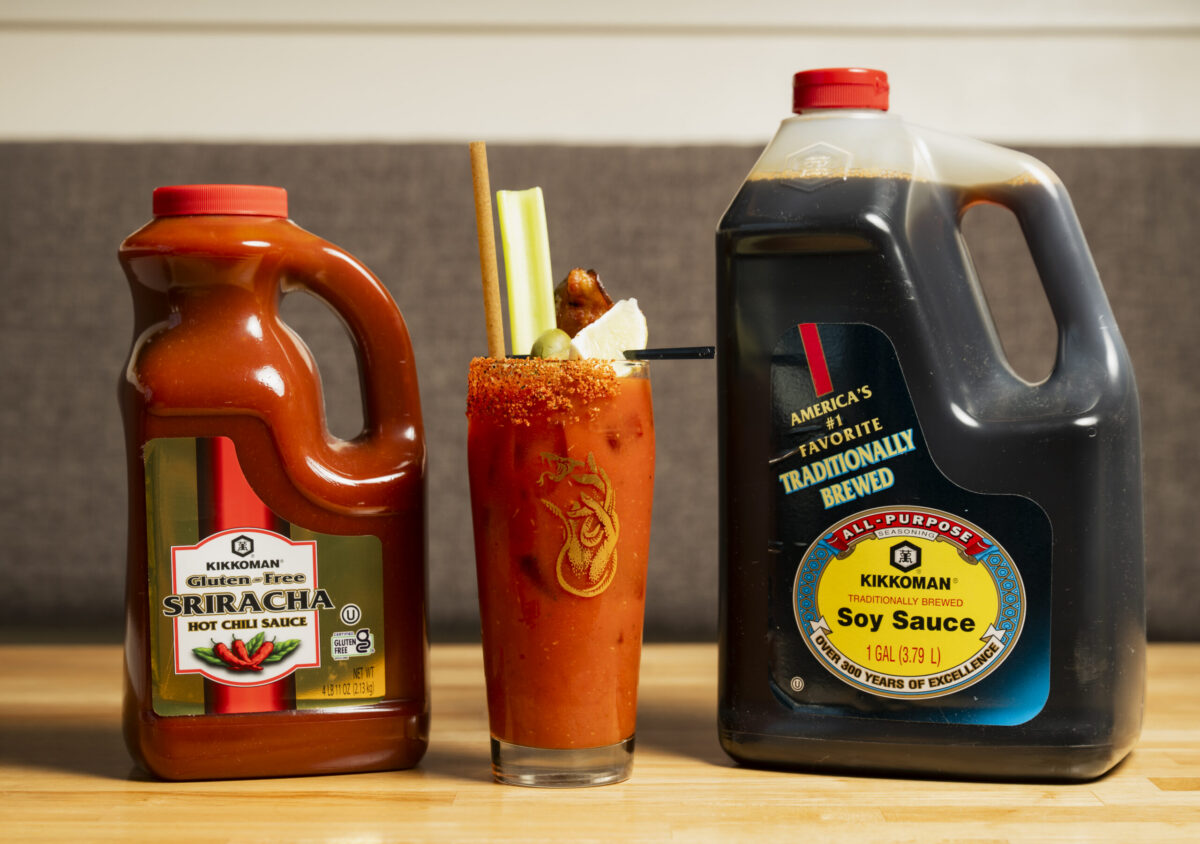 A jug of Kikkoman Sriracha sauce, a Bloody Mary cocktail with garnishes, and a jug of Kikkoman soy sauce are displayed on a wooden surface.