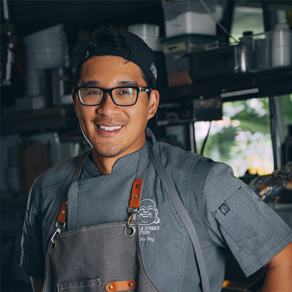 A man wearing glasses, a backwards cap, and a gray chef's uniform stands in a professional kitchen, smiling at the camera.