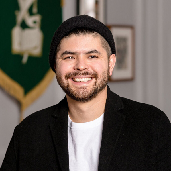 A man with a beard and mustache wearing a black beanie, black jacket, and white shirt smiles at the camera; a green and gold banner hangs in the background.