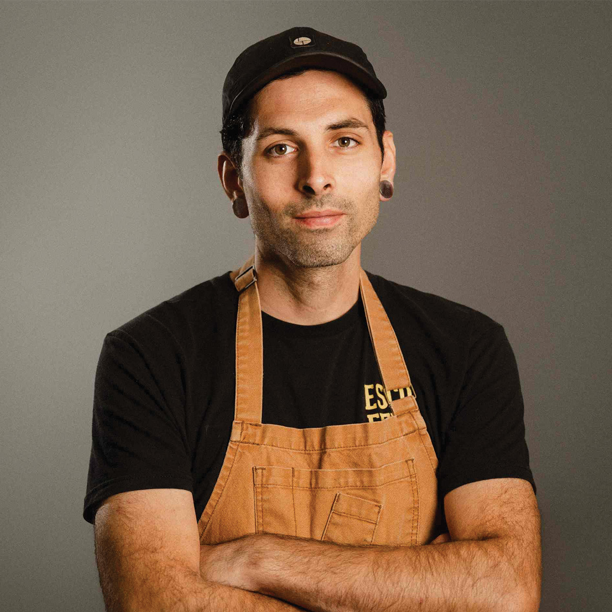A man wearing a black cap, black t-shirt, and tan apron stands with arms crossed against a plain background.