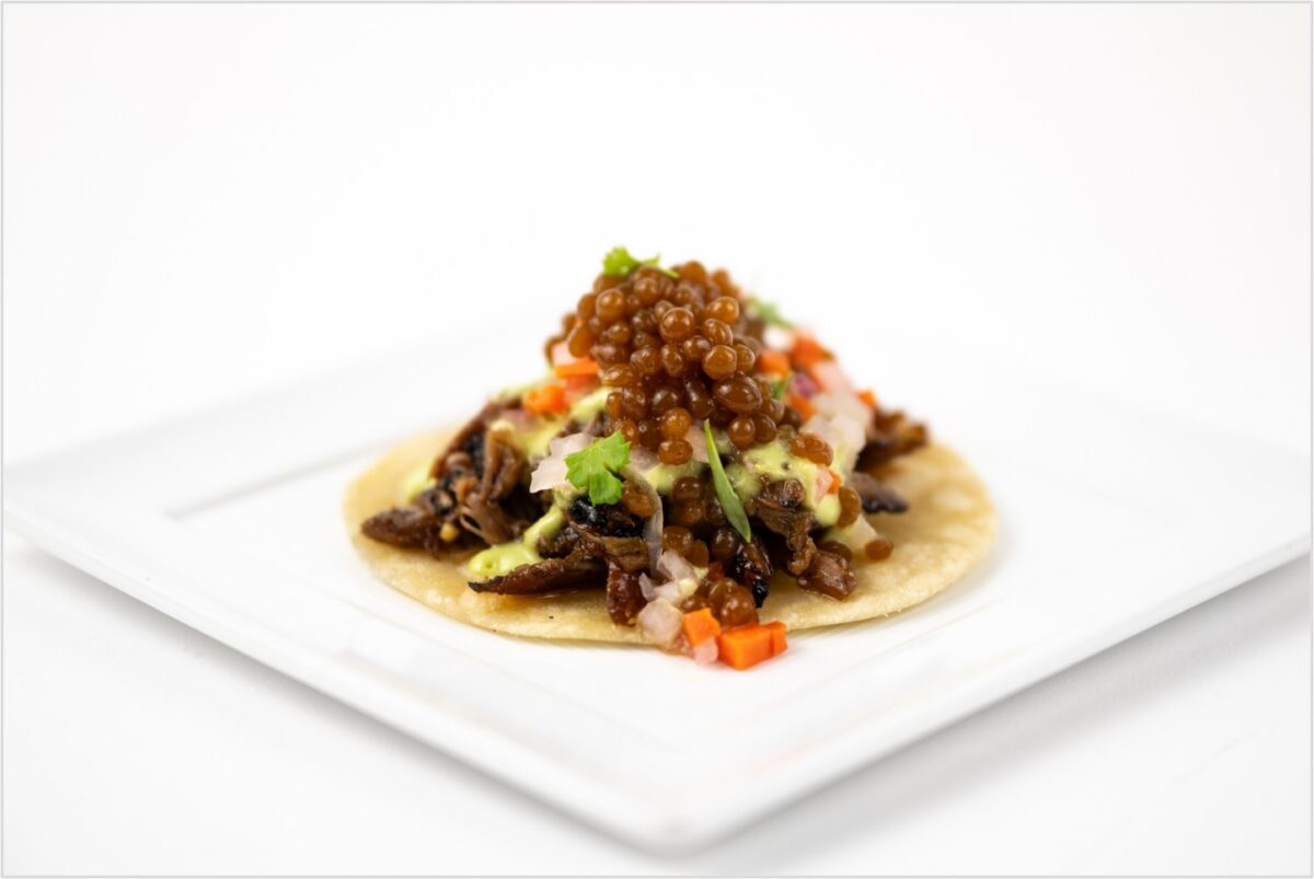 A small taco on a square white plate, topped with meat, chopped vegetables, cilantro, sauce, and a garnish of brown pearls.