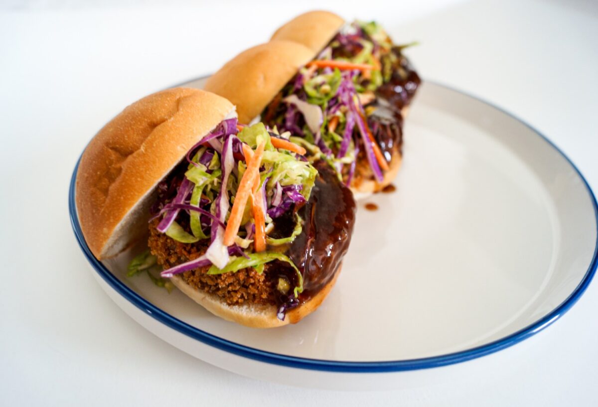 Two sandwiches on a plate, filled with breaded patty, barbecue sauce, and a colorful slaw of cabbage and carrots in a white room.