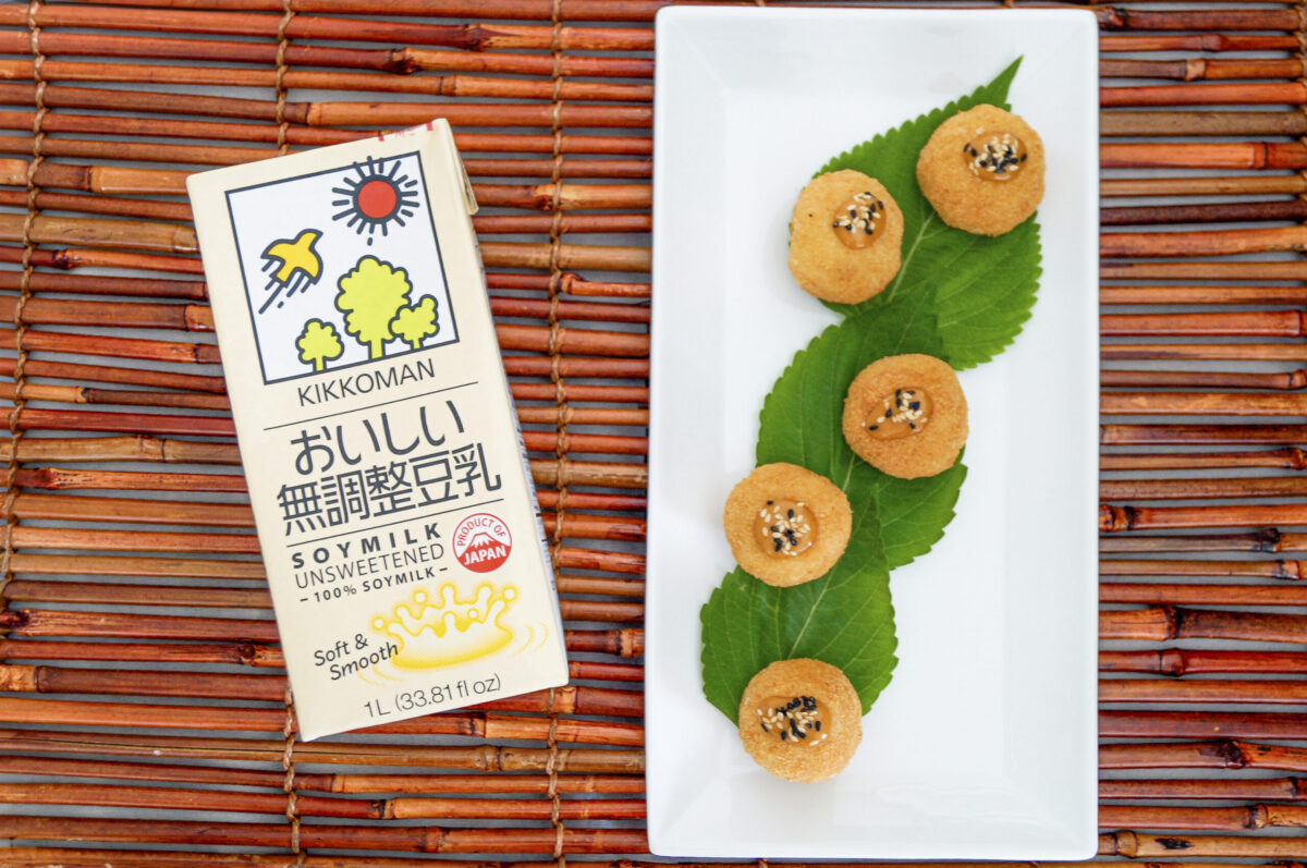 A carton of Kikkoman unsweetened soy milk sits next to five round snacks on green leaves, arranged on a bamboo mat.