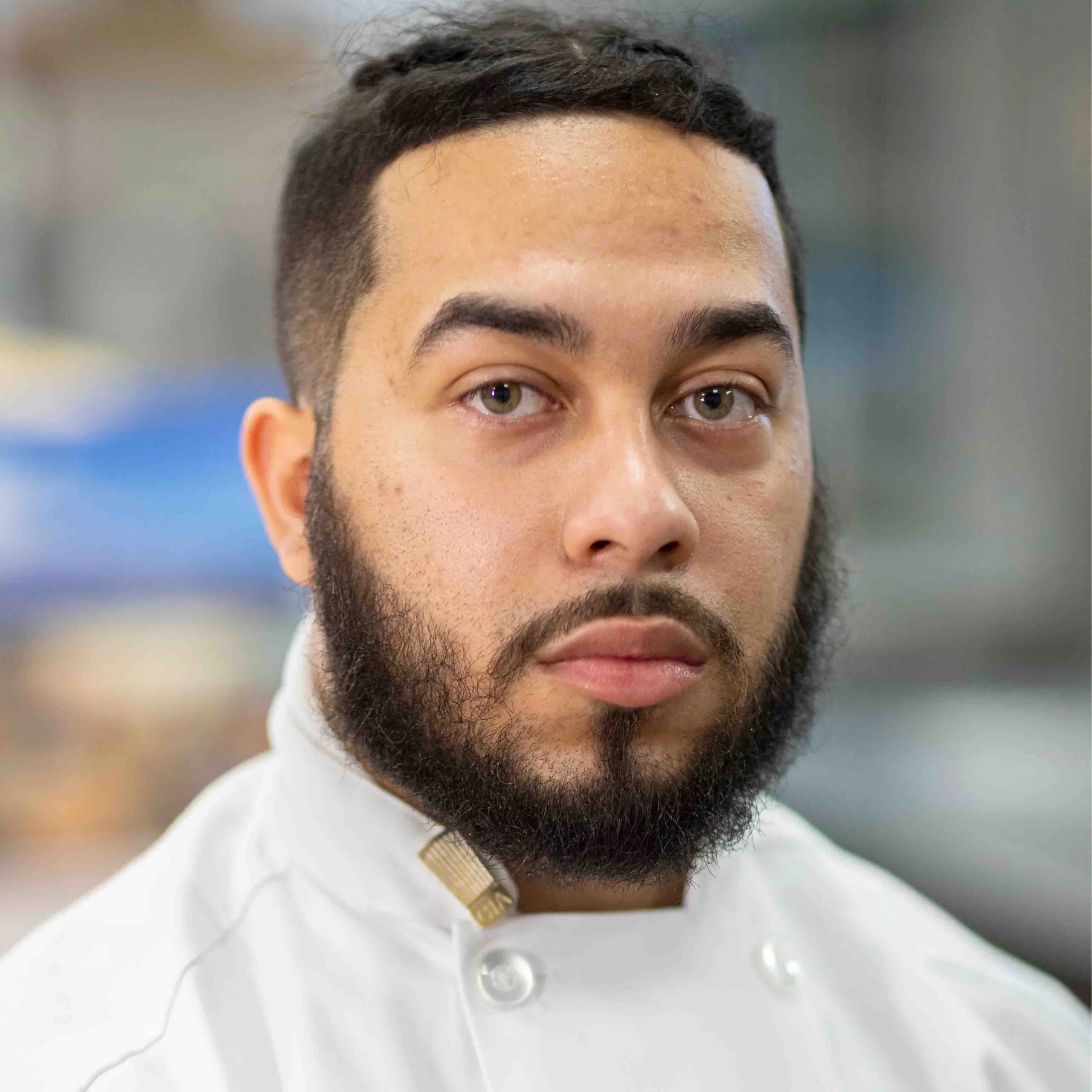 A man with a short beard and mustache wearing a white chef’s coat looks directly at the camera. The background is softly blurred.