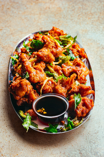 A plate of crispy fried chicken pieces garnished with herbs, served with a small bowl of dark dipping sauce on the side.