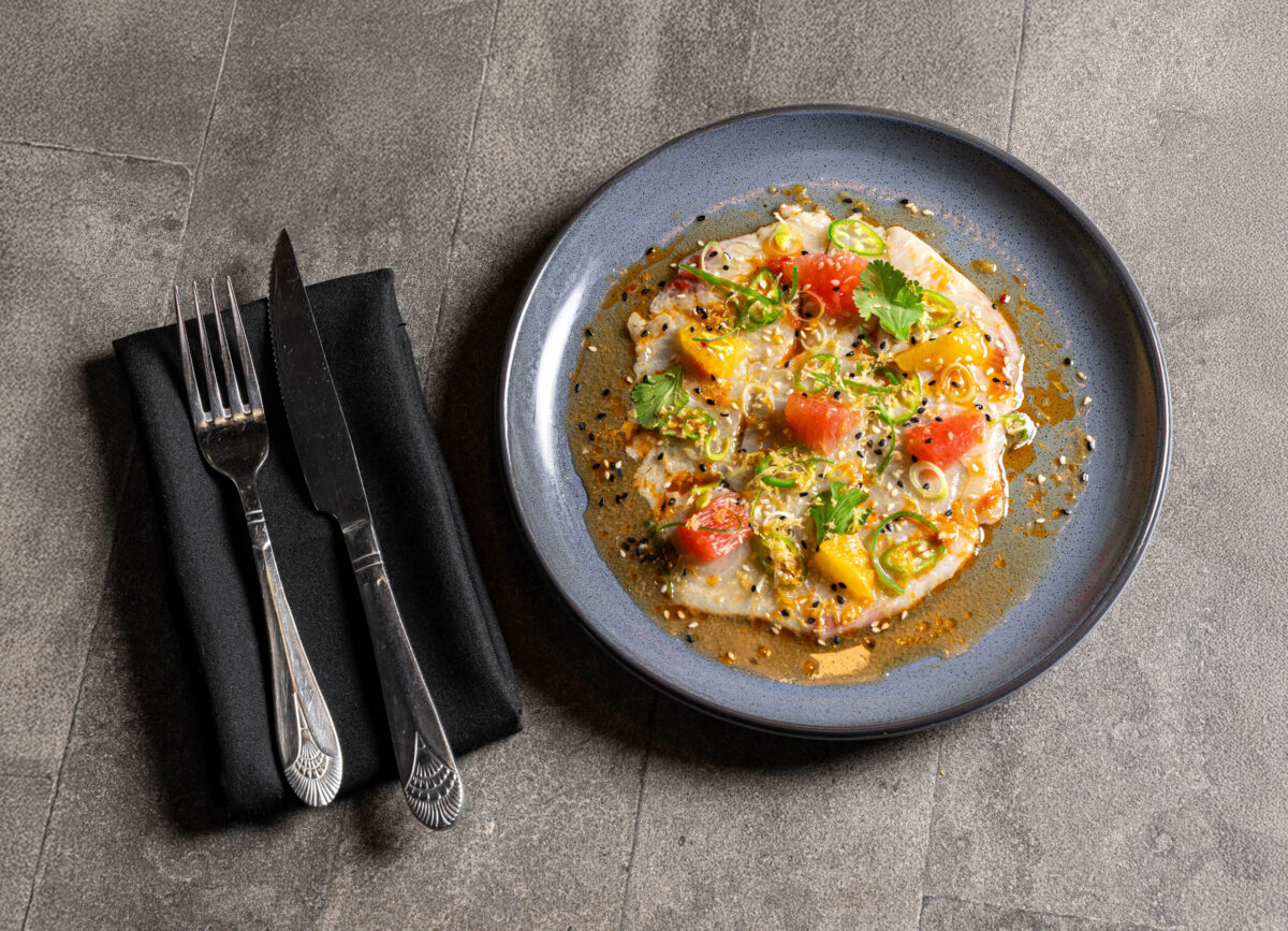 A plated dish of colorful sashimi garnished with herbs, sesame seeds, and sauce, next to a set of silverware on a black napkin atop a gray textured surface.