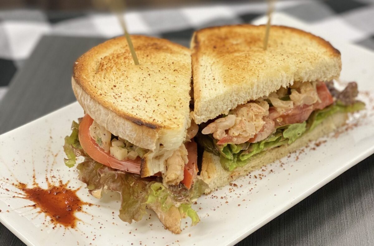 A toasted sandwich cut in half, filled with lettuce, tomato, bacon, and shredded chicken, served on a white plate with a drizzle of sauce.
